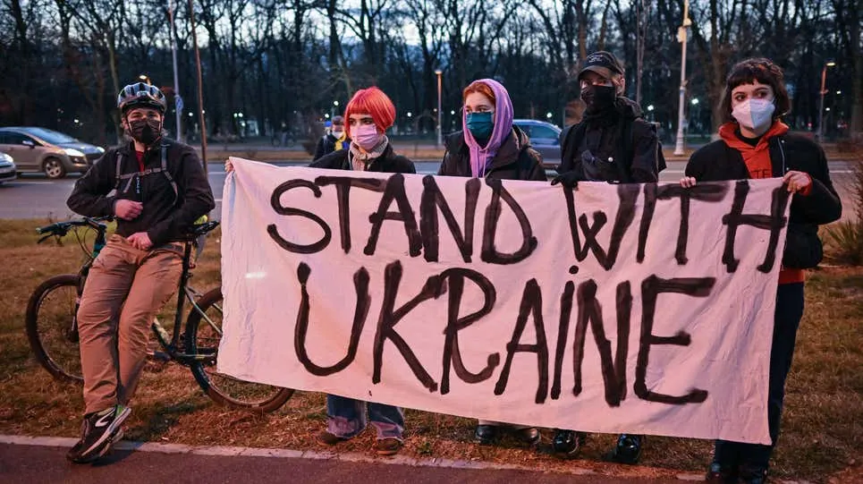 Five people wearing masks and standing outdoors hold a large sign that reads STAND WITH UKRAINE. There are trees and parked cars in the background, and one person has a bicycle.