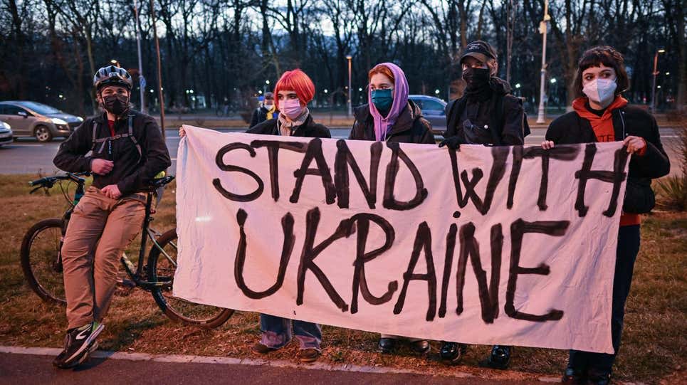 Five people wearing masks and standing outdoors hold a large sign that reads STAND WITH UKRAINE. There are trees and parked cars in the background, and one person has a bicycle.