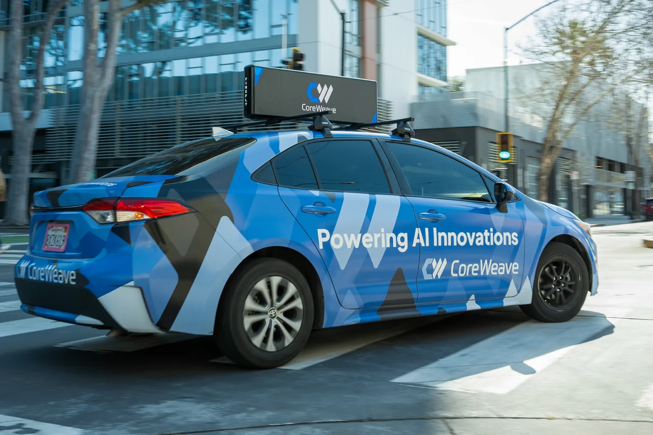 A blue CoreWeave-branded car with the slogan Powering All Innovations and a roof-mounted device drives through a city street with modern glass buildings in the background.