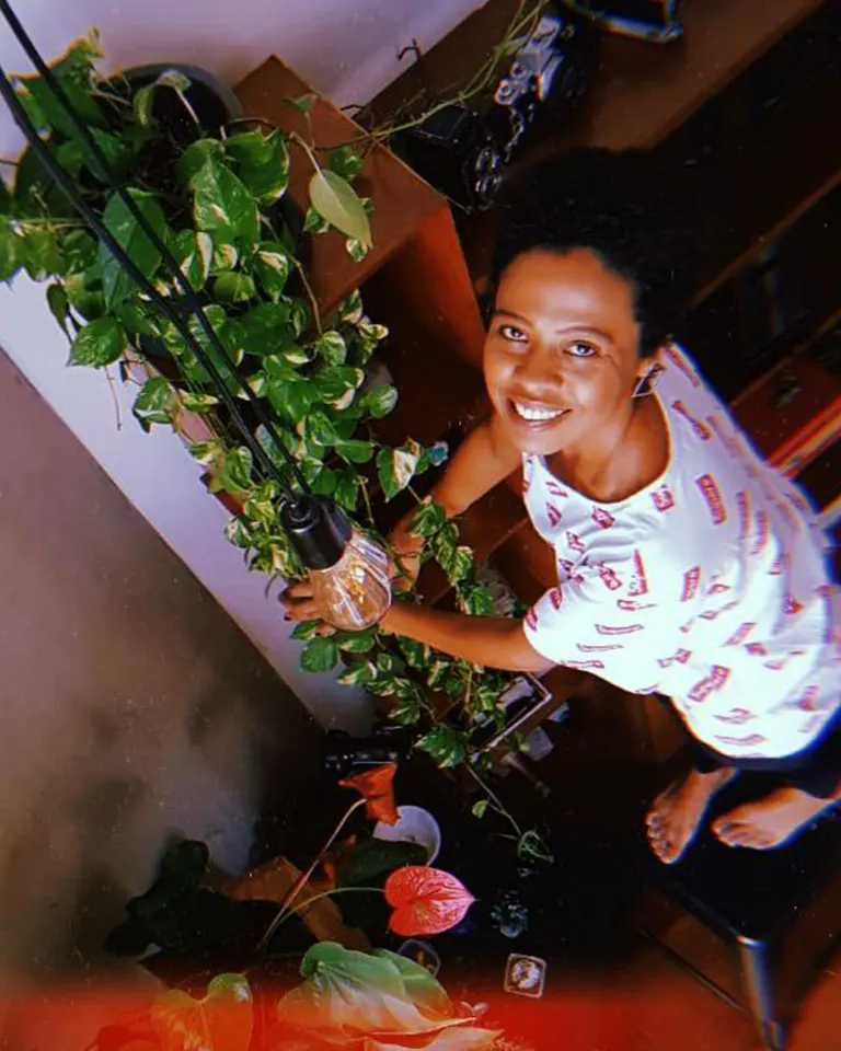 A person in a white shirt with colorful patterns smiles up at the camera while tending to a leafy green plant indoors, standing barefoot on a chair near a hanging lightbulb.
