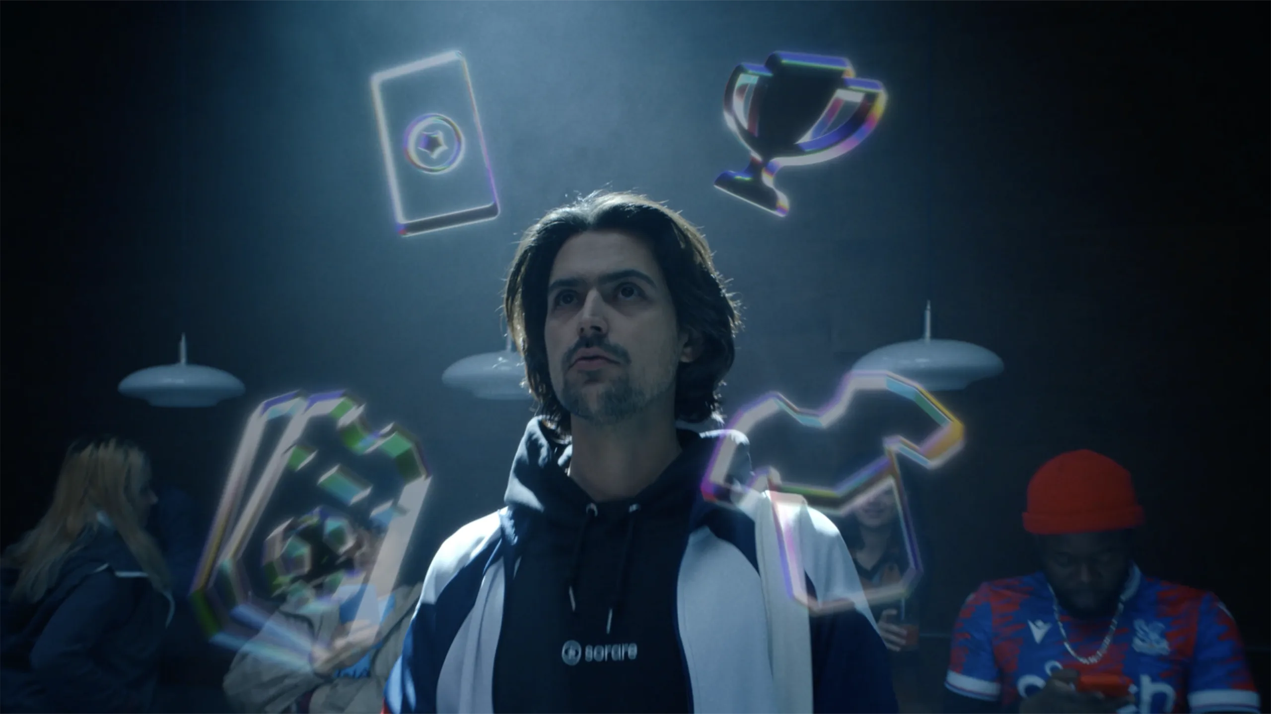 A man in a hoodie stands under a spotlight, looking up as floating holographic icons, including a trophy, t-shirt, card, and puzzle piece, hover above him. Blurred people and lights are in the background.