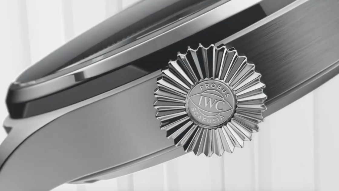 Close-up of a luxury wristwatch’s crown featuring a sunburst design and engraved with the words “PROBUS SCAFUSIA IWC.” The watch case is metallic and sleek with brushed and polished finishes.