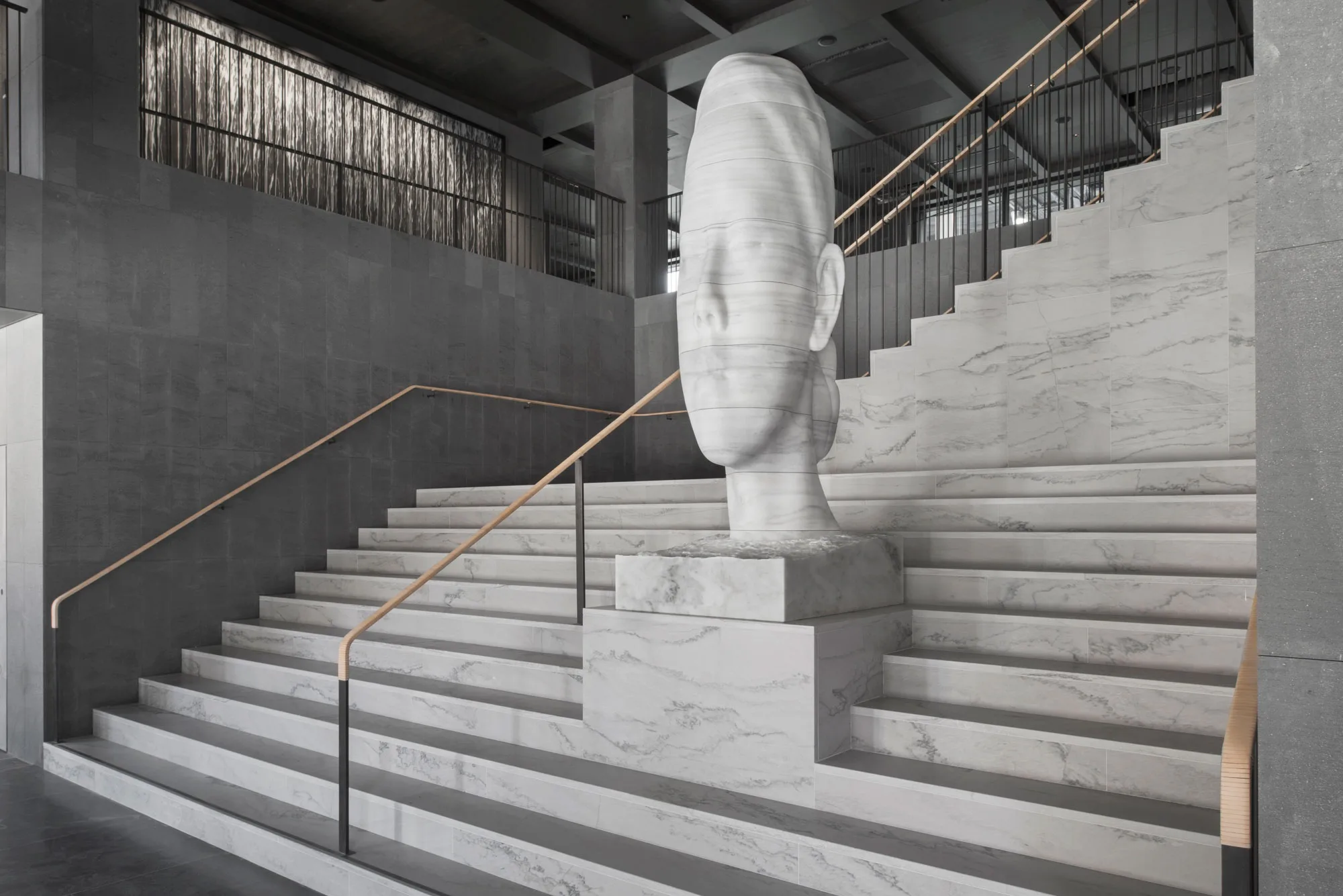 A large white marble sculpture of an abstract human head is positioned at the center of wide marble stairs with gold handrails in a modern, minimalist building interior.