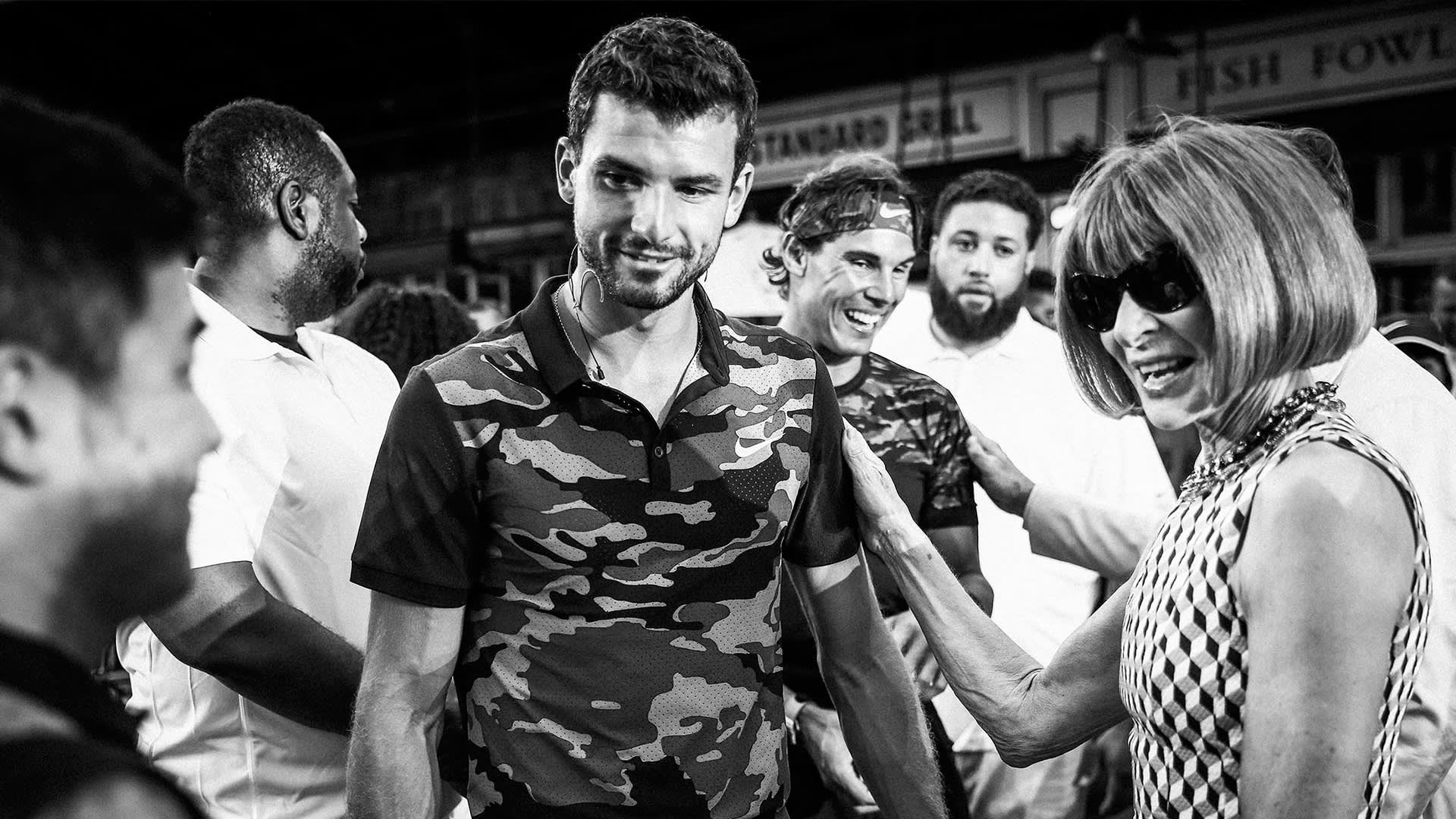 A group of people, including a smiling man in a camouflage shirt and a woman in sunglasses, interact and enjoy a lively moment indoors. The image is black and white.