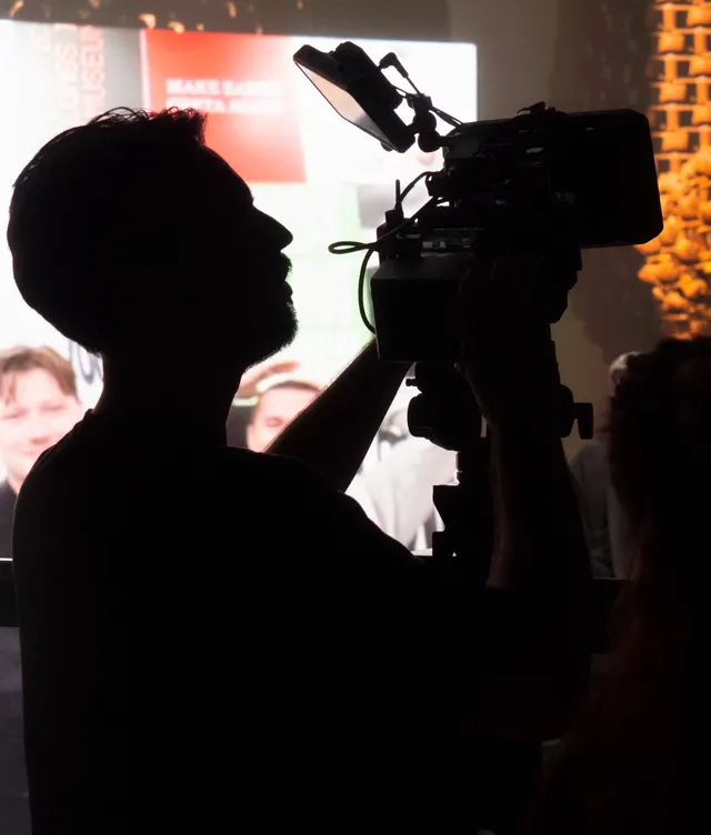 A silhouette of a camera operator filming an event, with a large, bright screen and blurry faces visible in the background. The camera and person are in sharp contrast due to the backlighting.