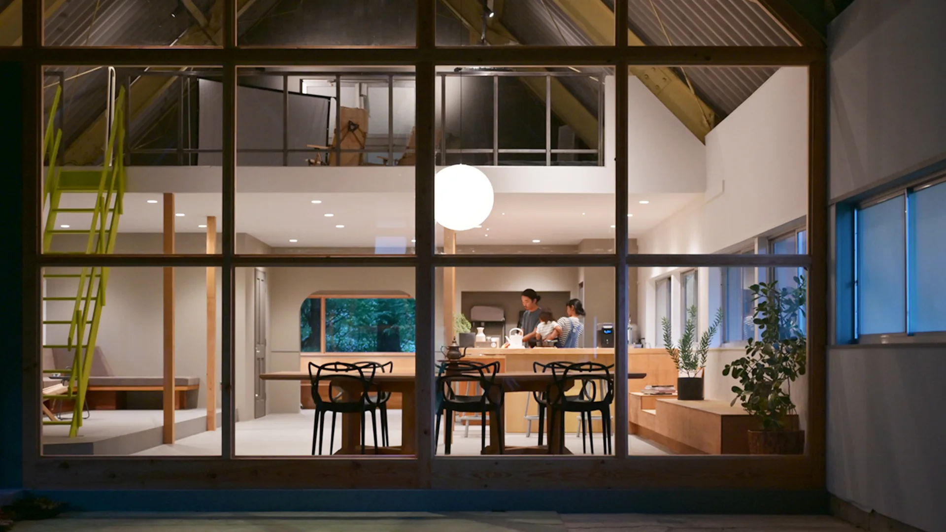 View through large windows into a modern, well-lit home; two people stand in the kitchen area, with plants, wooden furniture, and an open loft visible above.
