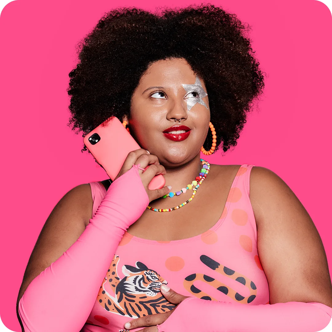 A woman with curly hair, wearing a colorful bead necklace, orange hoop earrings, and a pink outfit with bold patterns, holds a pink phone. She has silver makeup over one eye and stands against a bright pink background.
