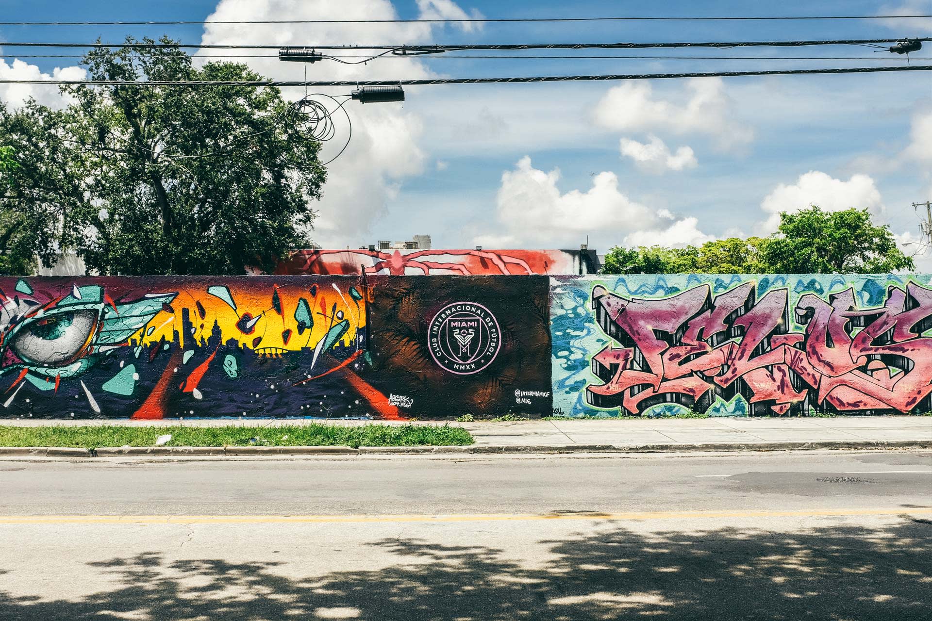 A colorful street mural on a long wall features an abstract eye, vibrant cityscape, stylized text, and the Inter Miami CF soccer logo at the center, with trees and a blue sky in the background.