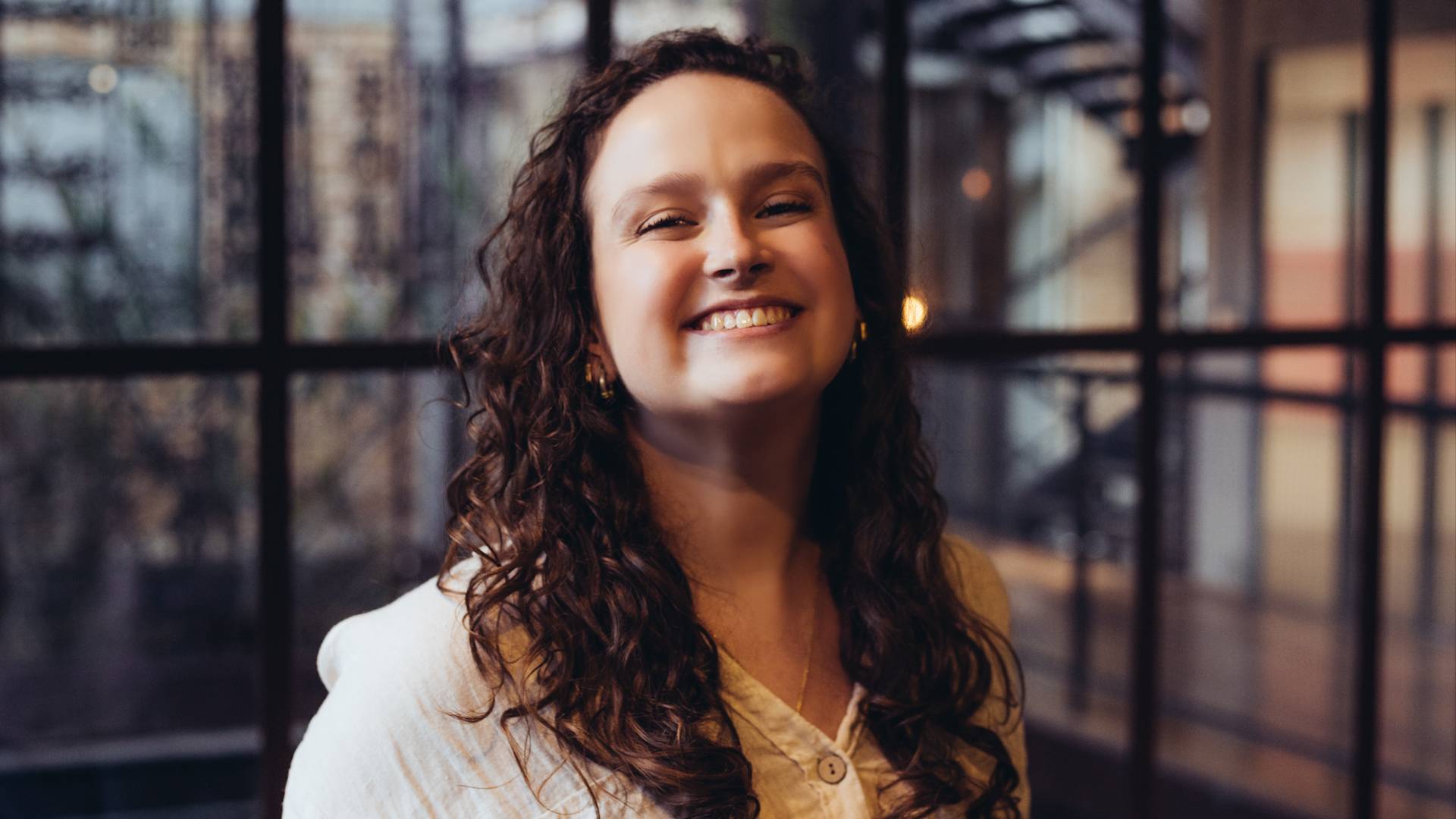 A woman with long curly hair, wearing a light-colored top, smiles while standing indoors near large windows with a blurred background of a modern building.
