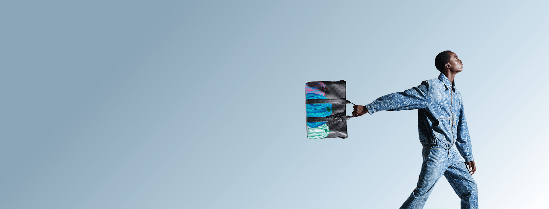 A person in a blue denim outfit walks confidently, holding a colorful bag outstretched to the side, against a gradient light blue background.