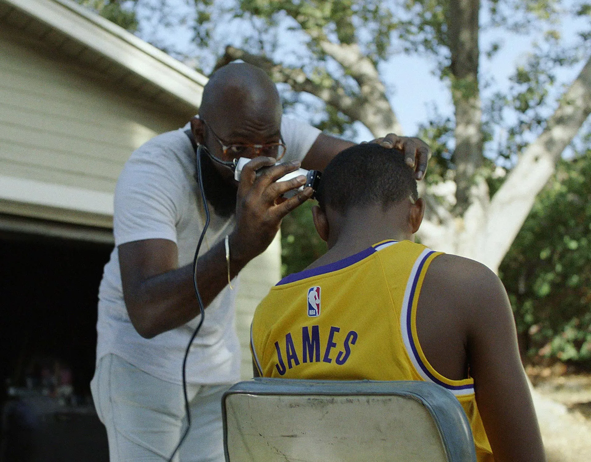 A man gives a haircut to a boy wearing a yellow basketball jersey with James on the back, outdoors near a garage and trees on a sunny day.
