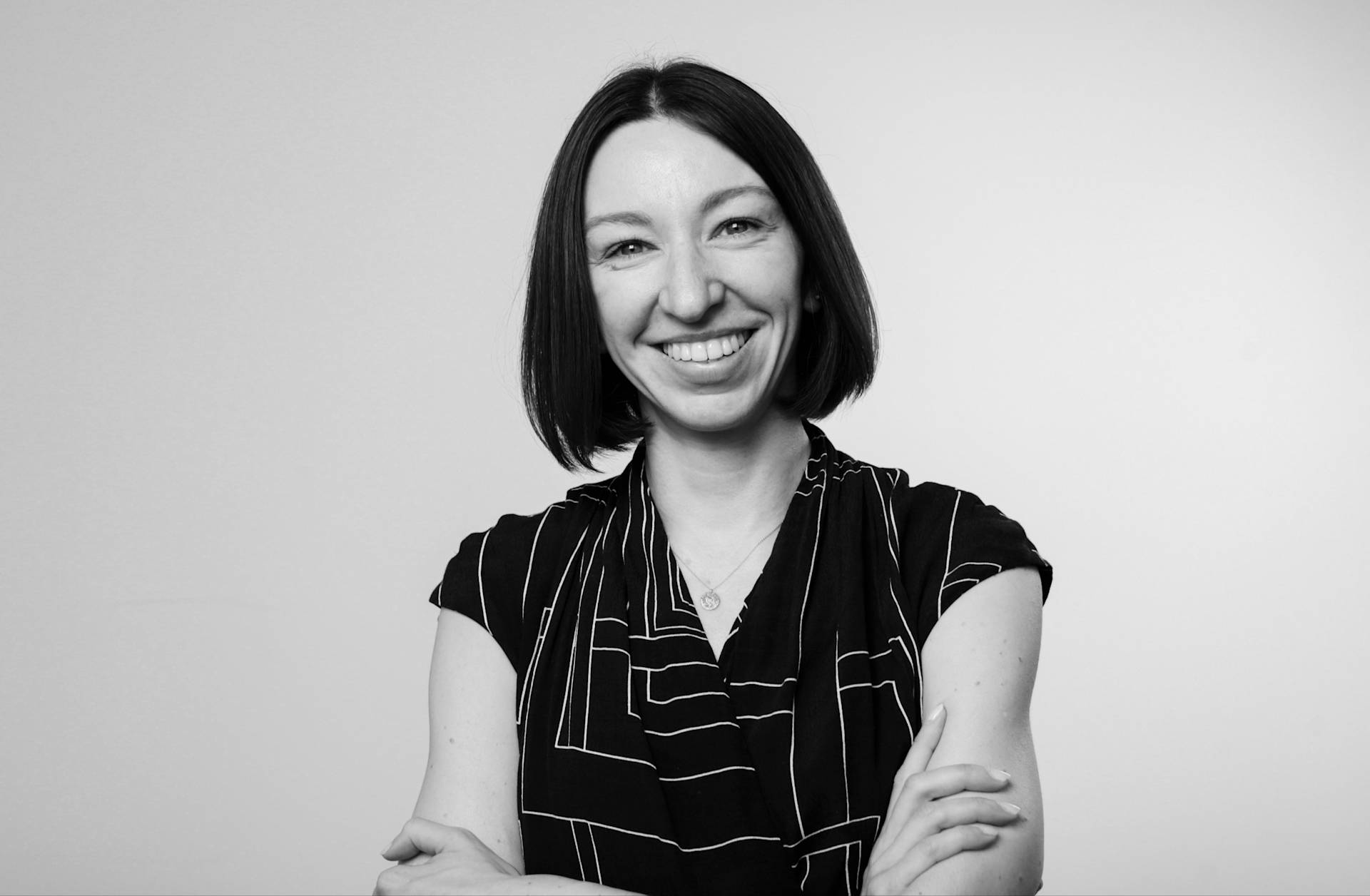 Black and white photo of a woman with straight dark hair, smiling and standing with arms crossed. She wears a patterned blouse and stands against a plain, light background.