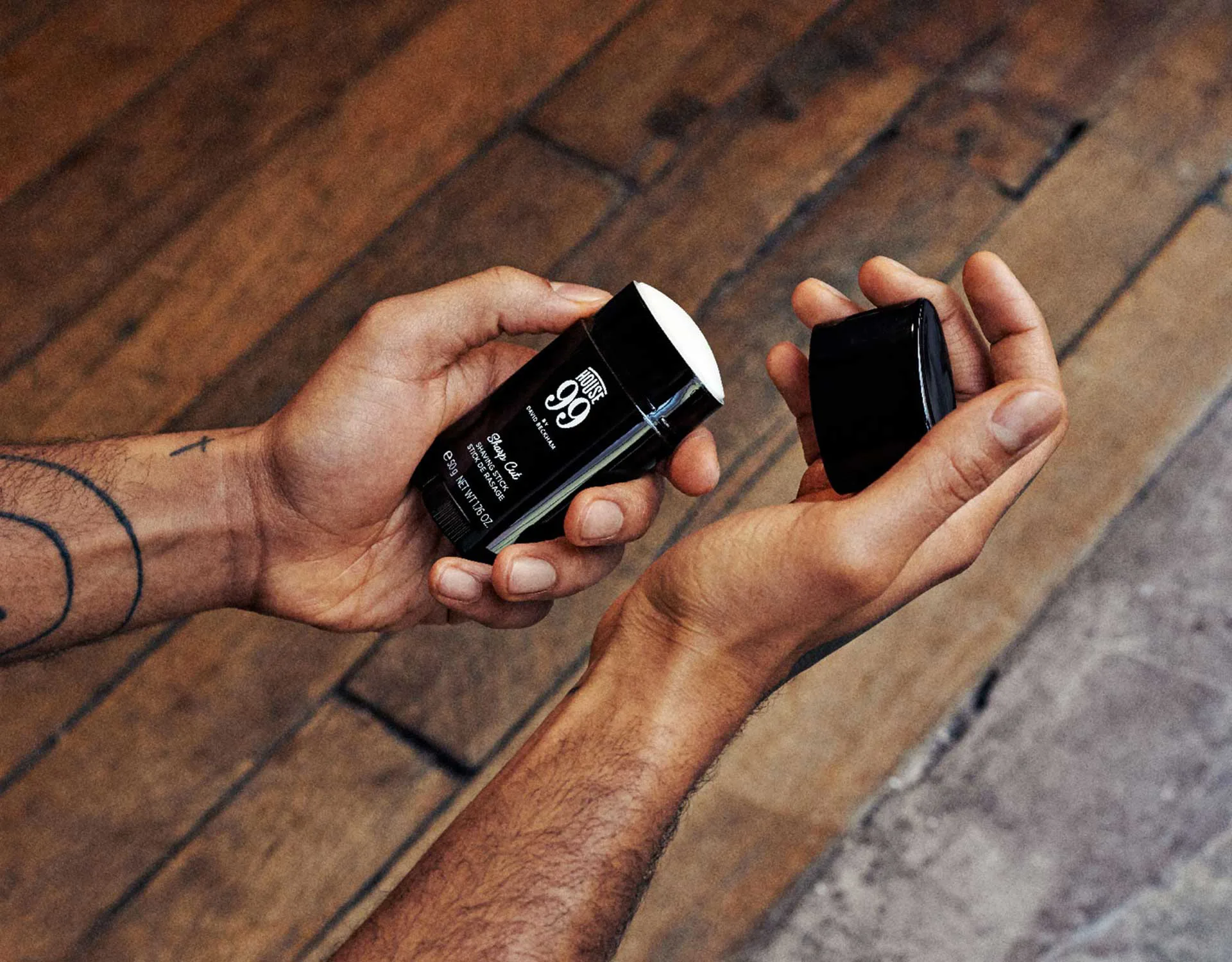A person with tattoos holds an open stick of 96-hour deodorant in one hand and the cap in the other, over a wooden floor background.