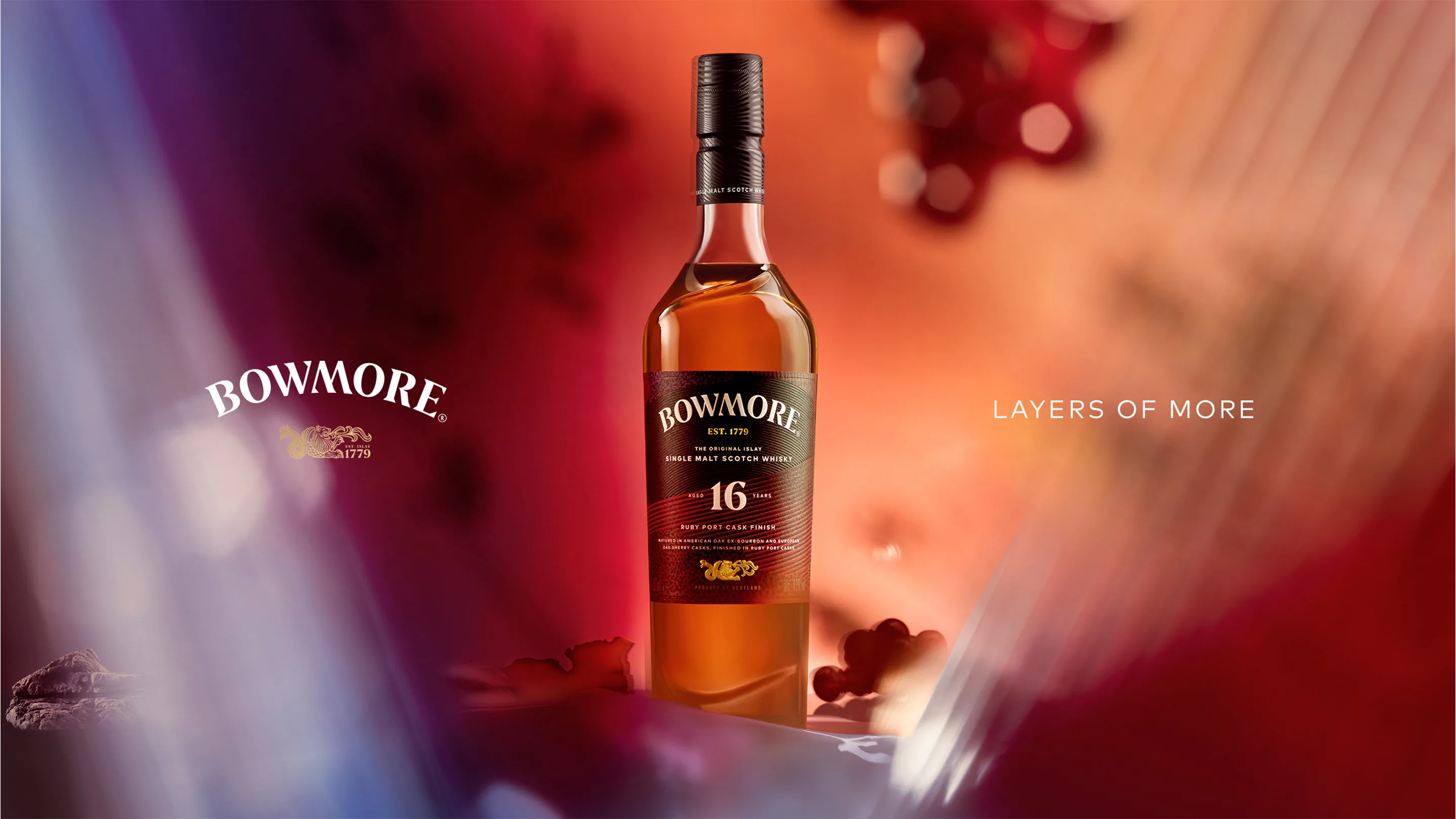 A bottle of Bowmore 16 Year Old Islay Single Malt Scotch Whisky stands upright against a warm, blurred background with red and orange hues. The words LAYERS OF MORE appear to the right of the bottle.