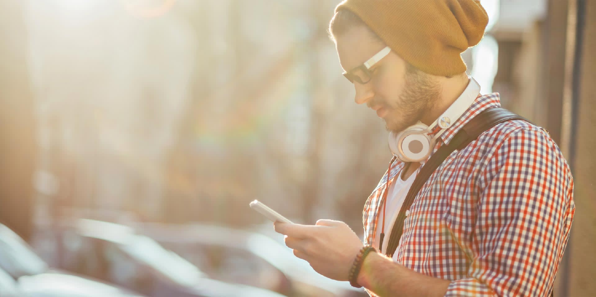 A young man wearing a beanie, sunglasses, and headphones around his neck stands outdoors, looking at his smartphone. Sunlight creates a soft, warm glow in the background.