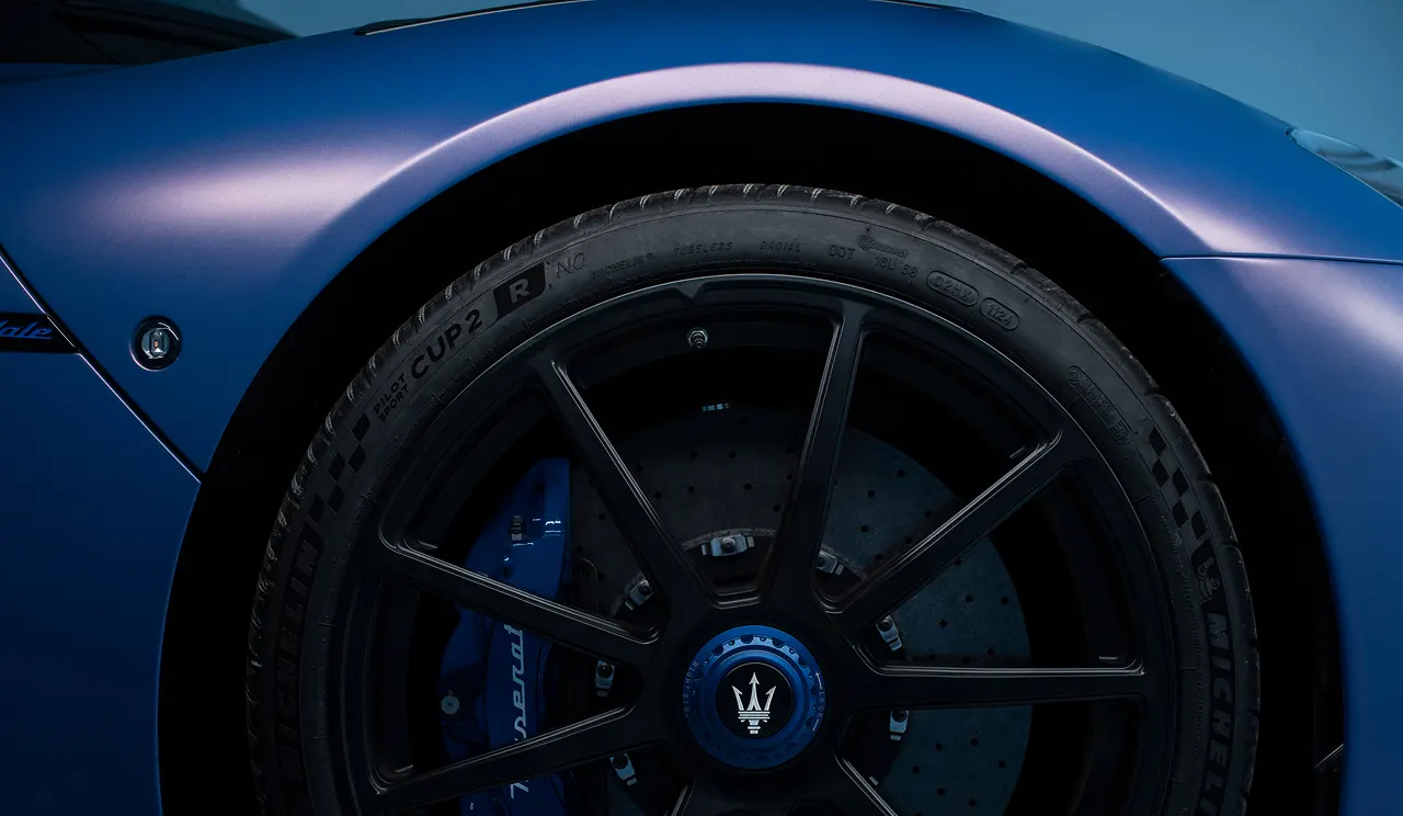 Close-up of a matte blue Maserati sports cars front wheel, featuring a black rim with the Maserati logo, visible brake disc, and blue brake caliper.