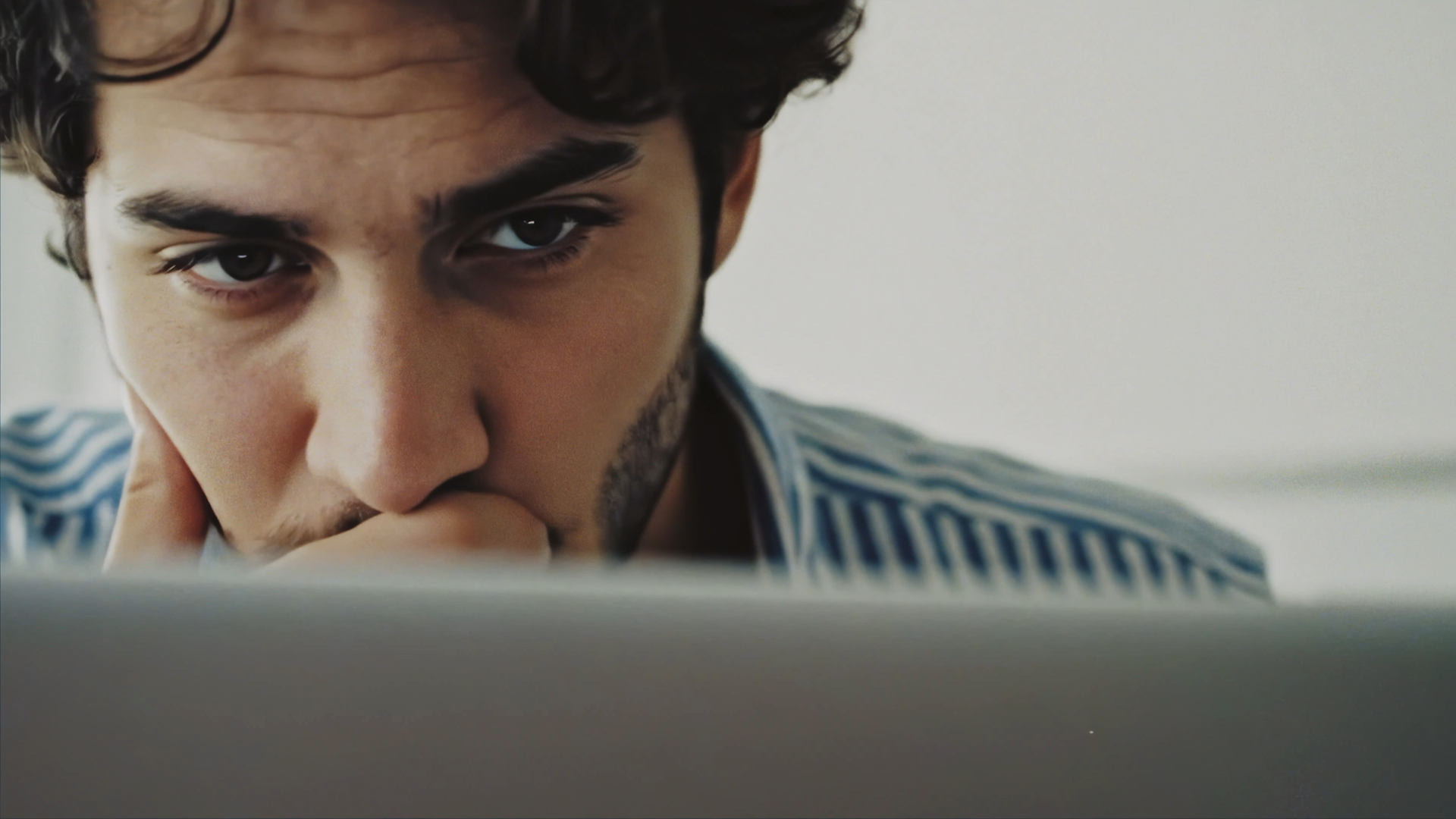 A young man with dark hair and a beard stares intently at a laptop screen, resting his chin on his hand while wearing a blue and white striped shirt. The mood appears focused and contemplative.