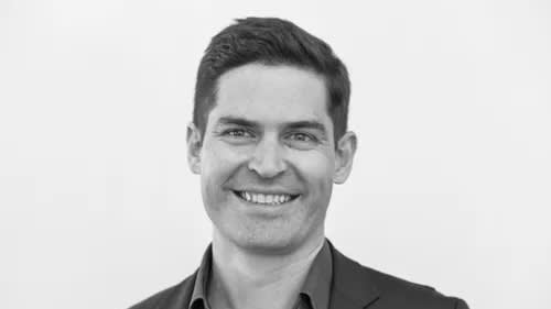 Black-and-white photo of a man with short dark hair, smiling. He is wearing a dark jacket and light shirt, standing against a plain white background.