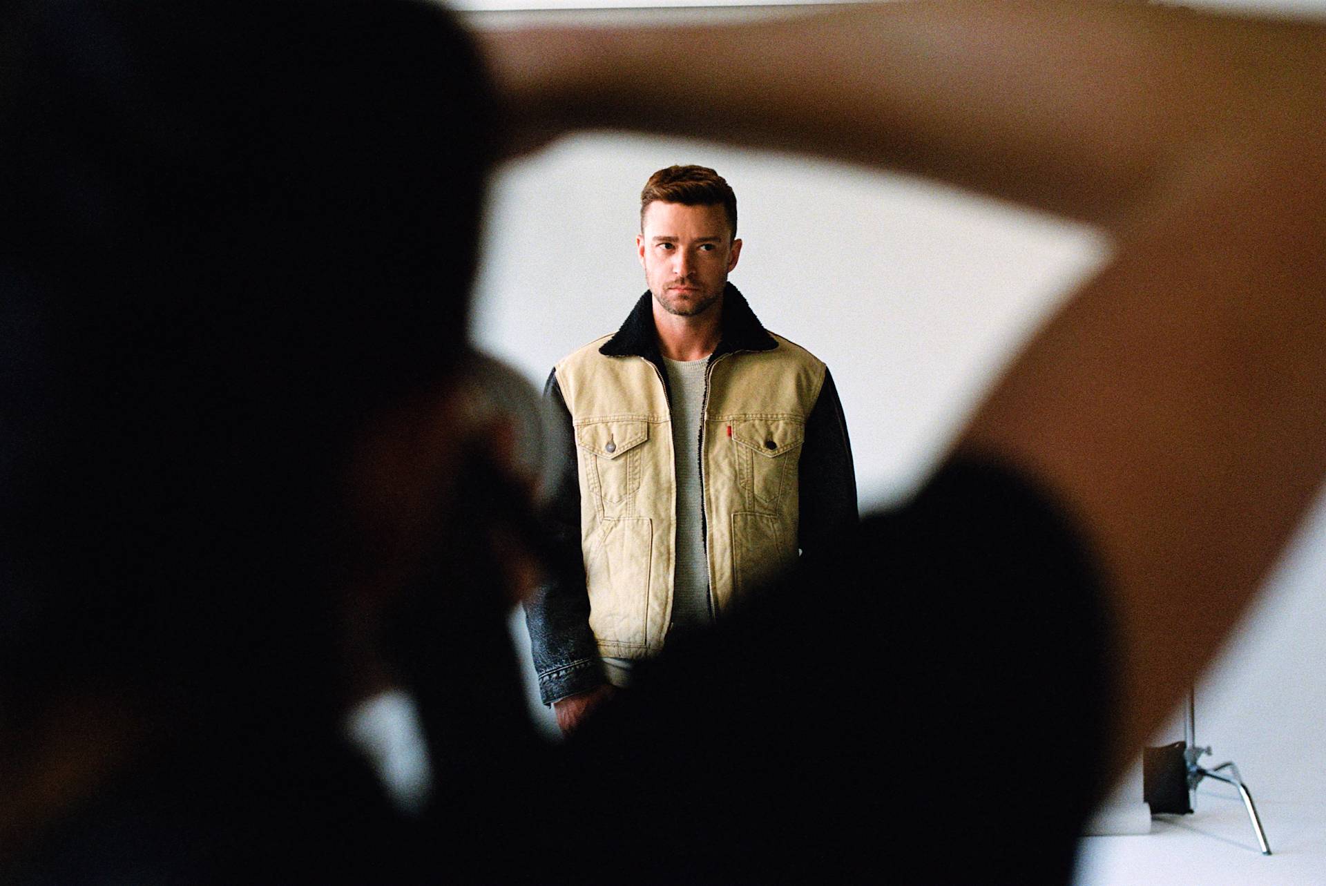A man in a beige and black jacket stands against a plain white background, looking serious. A photographers arm and camera are visible in the foreground, slightly out of focus.
