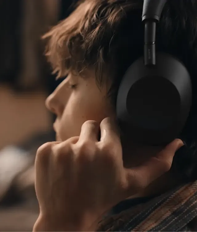 A person with brown hair wears large black over-ear headphones and touches the ear cup with their fingers, appearing to listen intently. The background is softly blurred.