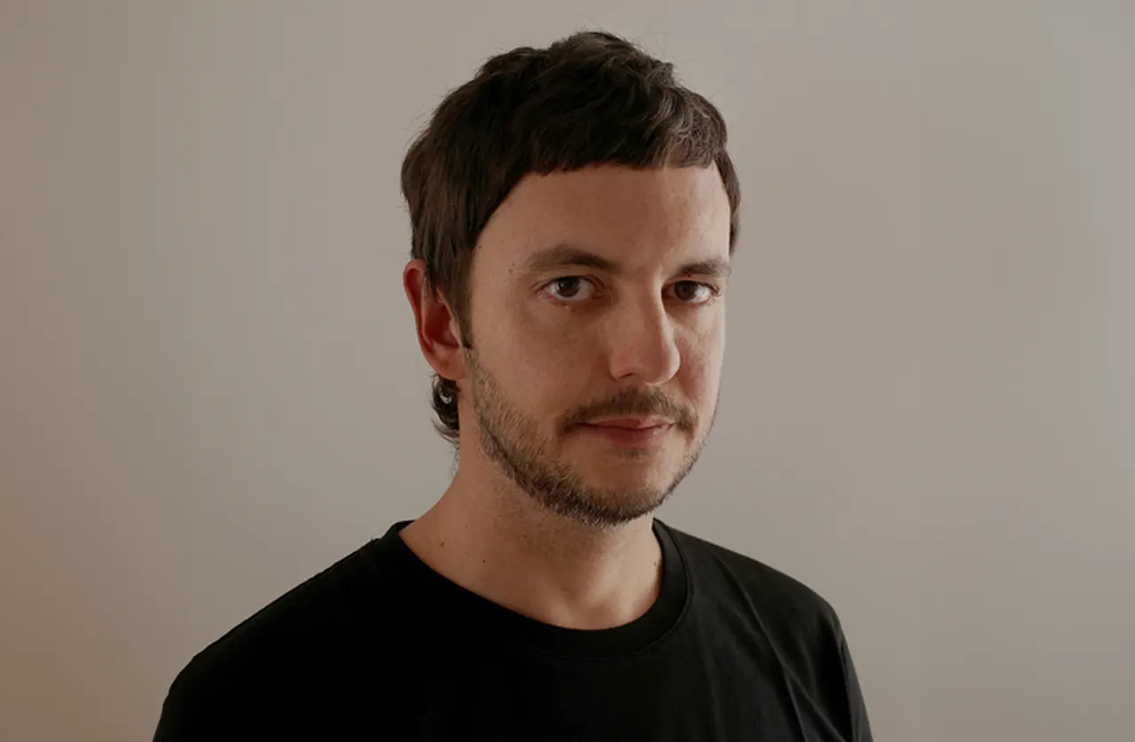 A man with short brown hair, a trimmed beard, and a neutral expression faces the camera in front of a plain light-colored wall. He is wearing a black t-shirt.