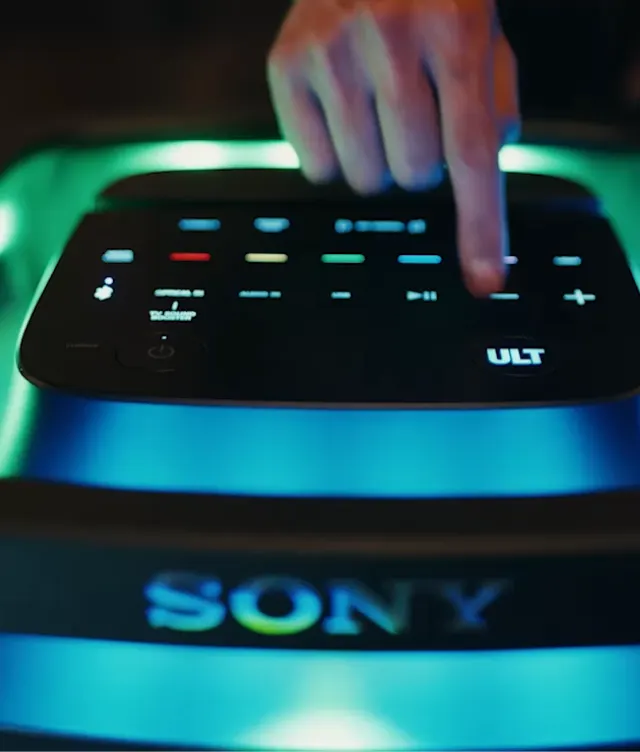 A hand presses a glowing button on top of a Sony speaker system with illuminated touch controls and the Sony logo visible in blue light.