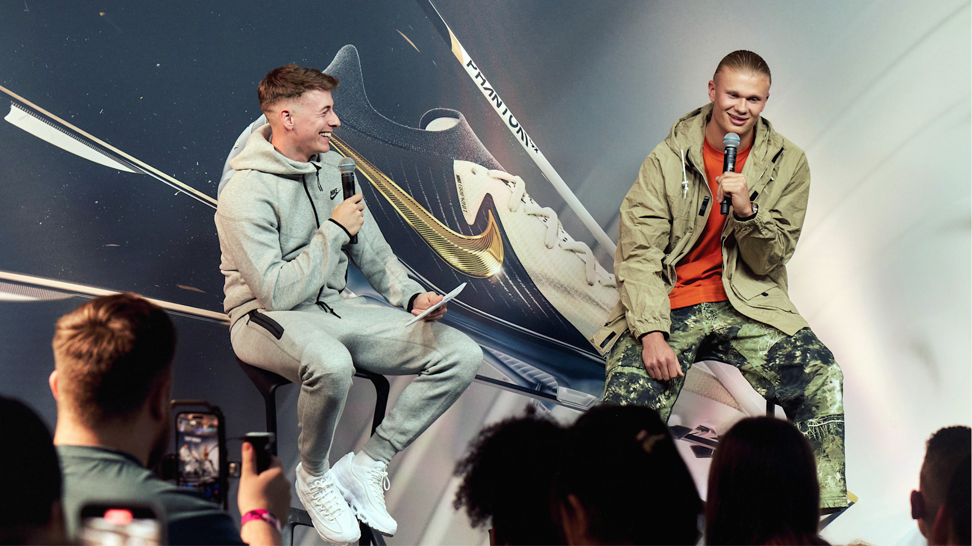 Two men sit on stools speaking into microphones at an event. One wears a gray tracksuit, the other wears a beige jacket and patterned pants. They are in front of a large display featuring a Nike Phantom soccer shoe. Audience members watch.