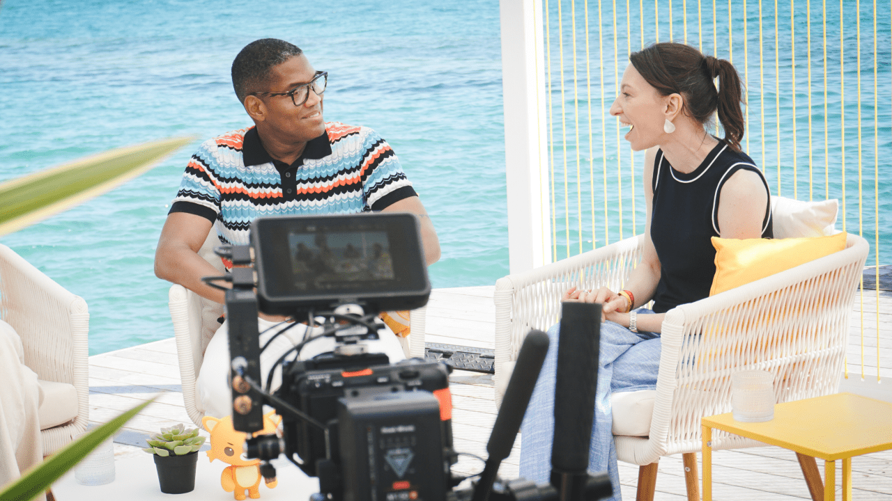 Tye Comer and a Gemma Redgrave sitting in white wicker chairs on a wooden deck overlooking the ocean. Tye , on the left, is wearing a striped polo shirt and glasses, looking at the Gemma and smiling. Gemma, on the right, is wearing a dark top and light blue pants, looking at the man and laughing. A camera is visible in the foreground.