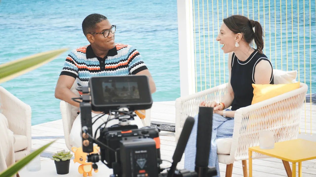 Tye Comer and a Gemma Redgrave sitting in white wicker chairs on a wooden deck overlooking the ocean. Tye , on the left, is wearing a striped polo shirt and glasses, looking at the Gemma and smiling. Gemma, on the right, is wearing a dark top and light blue pants, looking at the man and laughing. A camera is visible in the foreground.