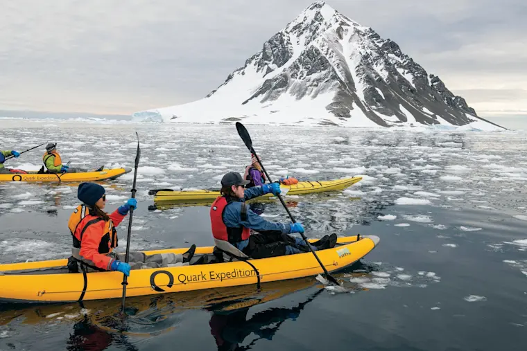 Kayakkers van Quark Expeditions peddelen tussen drijvende ijsschollen in Antarctic waters met een imposante sneeuwbedekte berg op de achtergrond.