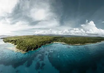 Luchtfoto van een tropisch eiland met dicht regenwoud, omgeven door turquoise zee en wit zandstrand, onder een dramatische bewolkte hemel.