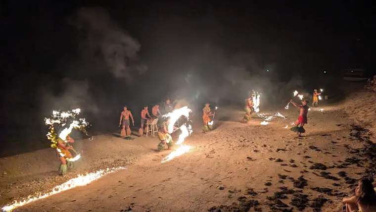 Een groep mensen voert 's nachts een spectaculaire vuurshow op met brandende fakkels op een verlicht zandstrand, waarbij vlammen hoog oplaaien tegen de donkere nacht.