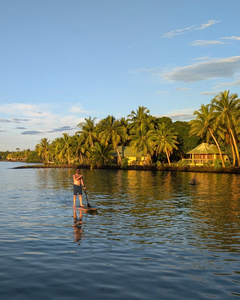 Een serene rivierlandschap met palmbomen aan beide oevers waar een persoon op een stand-up paddleboard over het kalme water vaart, onder een mooie blauwe hemel met wolken.