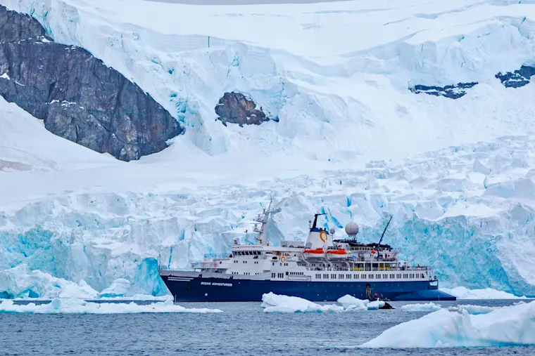 Een onderzoeksschip navigeert door imposante ijsbergen en gletsjers in de arctische wateren van Antarctica, omgeven door een adembenemend landschap van blauw ijs en rotsen.