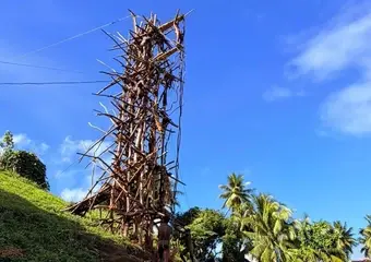 Een toerist fotografeert een indrukwekkende houten constructietoren op een groene heuvel met palmbomen in een tropisch landschap onder blauwe hemel.