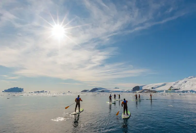 Een groep stand-up paddleboarders geniet van een zonnige dag op glasheldere poolwaters omgeven door gletsjers en ijsbergen in een arctisch landschap.
