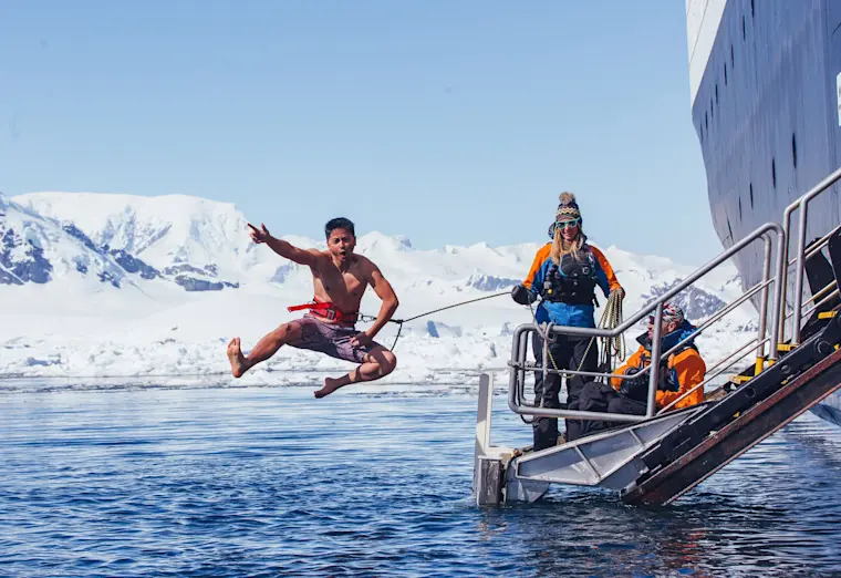 Een avontuurlijke man springt van een touwbruggetje in de ijskoude poolwateren voor de kust van Antarctica, terwijl toeristen vanuit de zodiac toekijken en besneeuwde bergtoppen op de achtergrond uitrijzen.