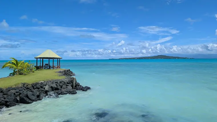 Een idyllisch tropisch strandparadijs met turquoise water, een elegante gazebo met rieten dak op een rotspunt, groene vegetatie en een eiland aan de horizon onder een wolkenloze blauwe hemel.