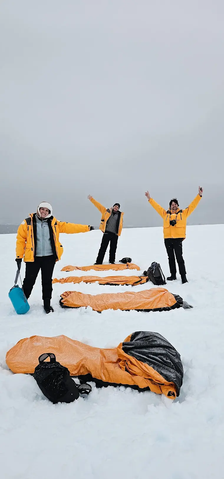 Een groep avonturiers in gele en zwarte winterkleding ligt en staat triomfantelijk in slaapzakken op een besneeuwde vlakte onder een grijze hemel.