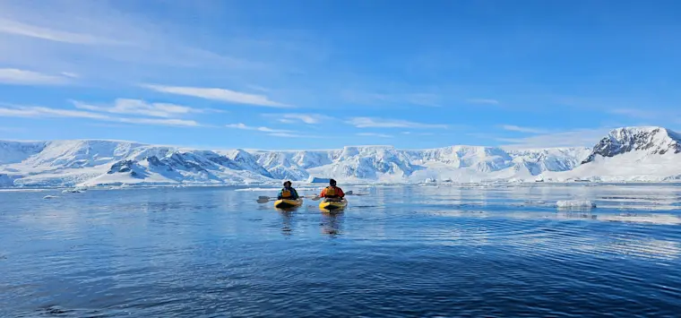 Twee kayakkers in gele zwemvesten paddelen over het kristalhelder blauwe water van een Arctische baai met imposante sneeuwbedekte bergen op de achtergrond.