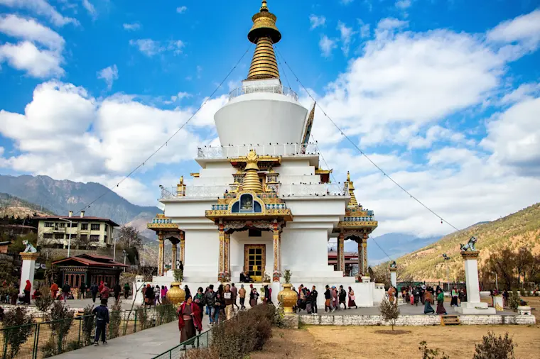 Een prachtige witte boeddhistische stupa met goudenDetailswerk omgeven door bergen, waar toeristen en lokale bezoekers bijeenkomen in een serene bergomgeving.