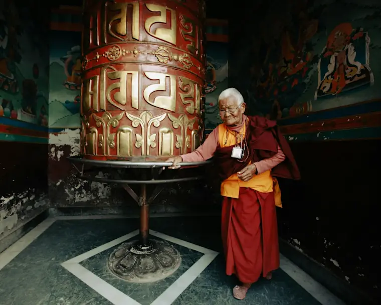 Een oudere Bhutanese monnik of gelovige die bij een grote, rode gebedsmolen (mani chokhor) staat.