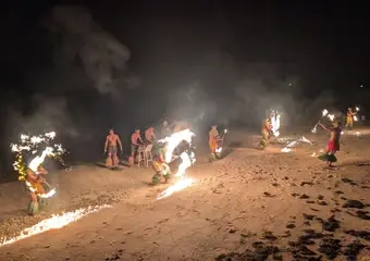 Een groep mensen voert 's nachts een spectaculaire vuurshow op met brandende fakkels op een zandstrand, wat een magische en avontuurlijke sfeer creëert.