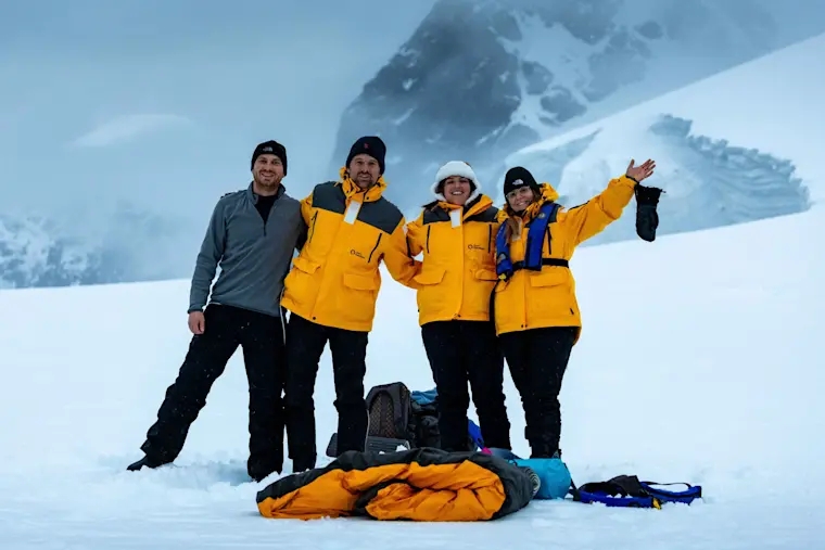 Een groep van vier enthousiaste expeditieleden in gele en zwarte winterjassen poseren samen op een besneeuwde bergflank met indrukwekkende besneeuwde pieken op de achtergrond, waarbij hun gele slaapzakken en berguitrusting zichtbaar zijn.