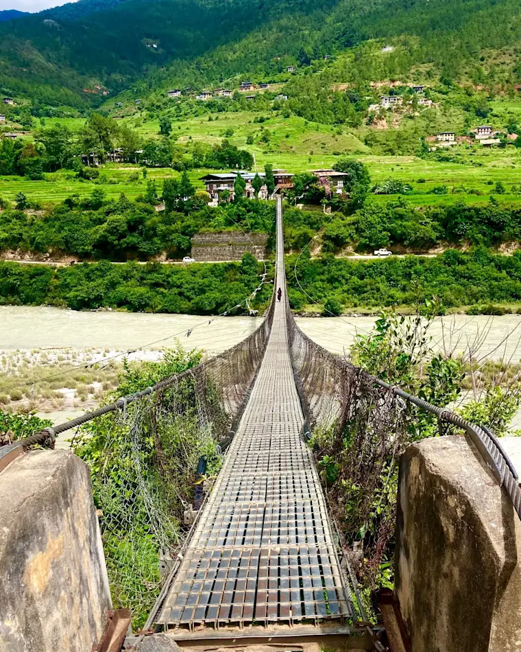 Een smal uitgehangen voetgangersbrug over een bergrivier in een groen dal met traditionele huizen en terrassen in wat lijkt te zijn Bhutan of de Himalaya-regio.