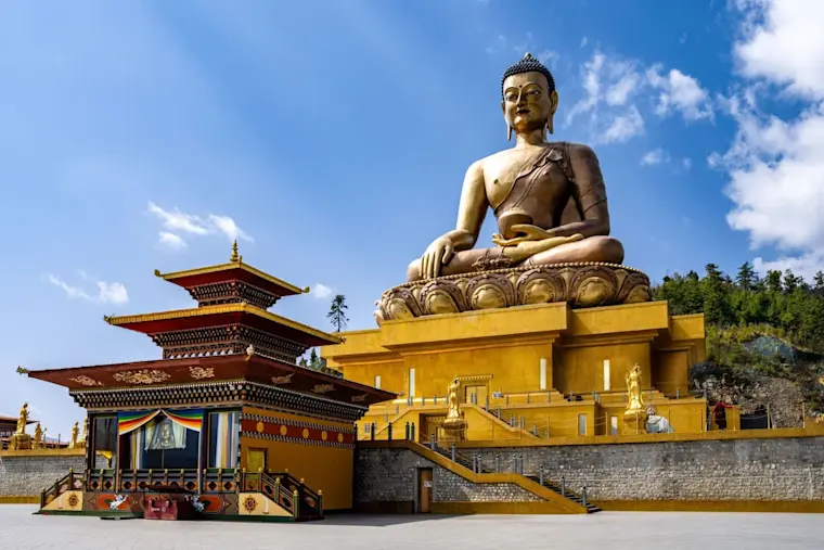 Een imposante gouden Boeddha-standbeeld zit in meditatie op een gele platform voor een traditioneel Bhutaans tempel met rode en gouden daken, omgeven door groene heuvels onder een blauwe hemel.