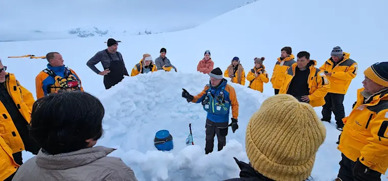 Een groep toeristen in gele winterjassen staat samen in een sneeuwlandschap waar een gids een groepsactiviteit of les geeft rond een sneeuwstructuur.