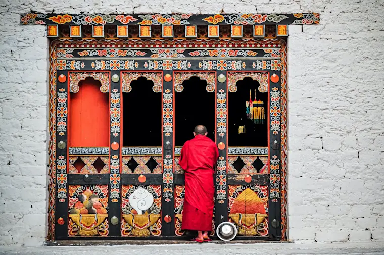 Een monnik in een rood gewaad die staat voor een rijkelijk gedecoreerde houten poort van een tempel vol prachtig traditioneel Bhutanese schilder- en snijwerk met levendige kleuren, symbolen en motieven op een ruwe, witgekalkte stenen muur.