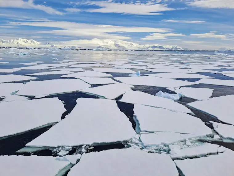 Adembenemend poollandschap met uitgestrekte pakijsformaties en donkere watergaten, omgeven door sneeuwwitte ijsschotsen en met besneeuwde bergen aan de horizon onder een partly bewolkte blauwe hemel.