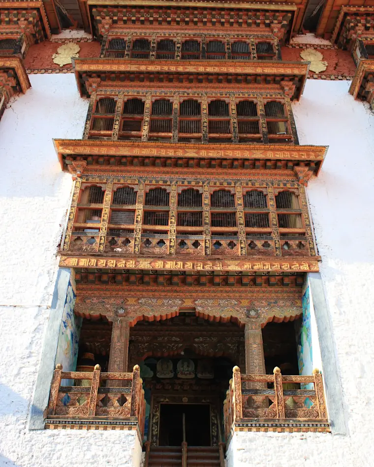 De metershoge, witgekalkte gevel van een Bhutanese tempel valt op door zijn rijk uitgesneden houten balustrades en uitstekende vensterpartijen.